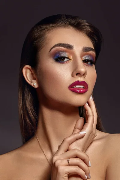 Naakte juwelier raakt haar gezicht, poseert op een grijze achtergrond. Luxe make-up. Kleurrijke oogschaduw, valse wimpers, glanzende rode lippen. Sluiten. — Stockfoto