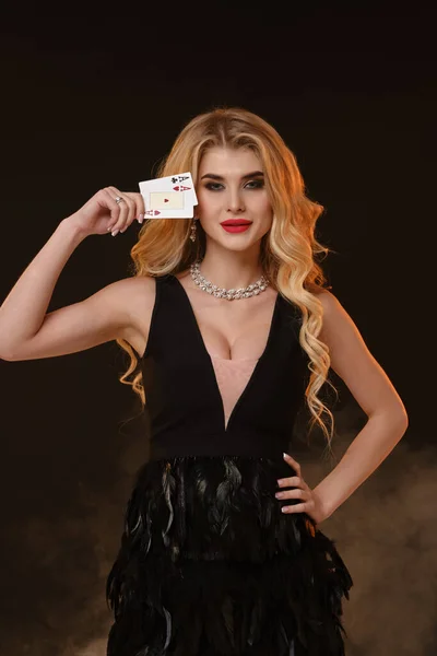 羽やネックレスと黒のドレスで見事な巻きブロンドの女性 明るいメイクアップ 黒のスモーキースタジオの背景にポーズを2枚のトランプを示しています ポーカー カジノ コピースペース — ストック写真