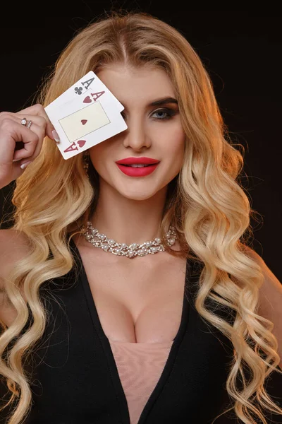 Gorgeous curly blonde model, bright make-up, in black stylish dress and necklace. She smiling, covered her eye by two playing cards, posing on black background. Poker, casino. Close-up, copy space