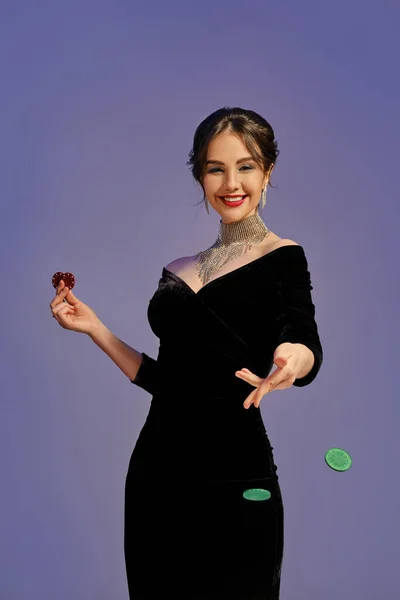ブルネットモデル 裸の肩 明るいメイクアップ 黒のドレスと光沢のあるジュエリーで 笑みを浮かべて 2つの赤いチップを保持し 緑のものを投げる 紫色の背景を持つ ポーカー カジノ — ストック写真