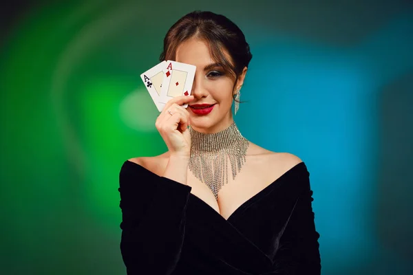 裸の肩と明るい化粧を持つブルネットの女の子 ブラックドレス 鮮やかなネックレスとイヤリングで カラフルな背景にポーズを2つの顔で彼女の目を覆った ポーカー カジノ — ストック写真