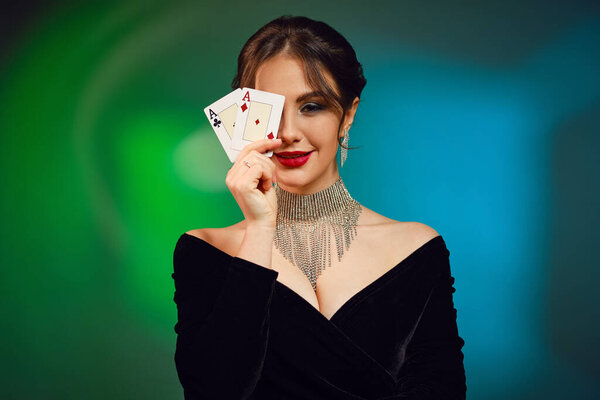 Brunette girl with bare shoulders and bright make-up. In black dress, brilliant necklace and earrings. Smiling, covered her eye by two aces, posing on colorful background. Poker, casino. Close-up