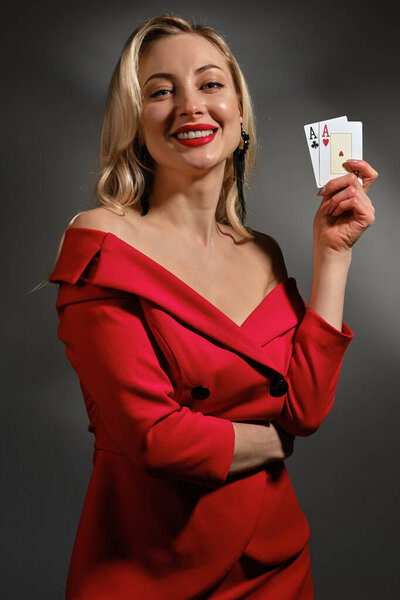 Cute blonde girl with bare shoulder, bright make-up, in red dress and black earrings. She is smiling, showing two playing cards, posing on gray background. Poker, casino. Close-up, copy space