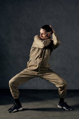Dövmeli, yüzlü, küpeli, sakallı genç, zarif bir sanatçı. Haki rengi tulum ve siyah spor ayakkabı giymişti. Gri arka planda dans ediyor. Dans salonu, hip-hop. Tam uzunluk, boşluğu kopyala