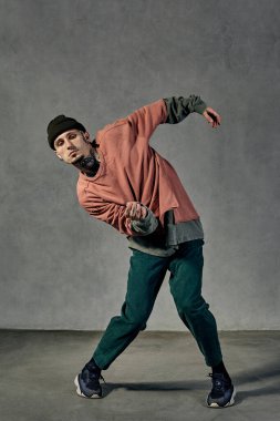 Dövmeli, yüzlü, küpeli, sakallı güçlü bir erkek. Şapkalı, renkli kazaklı, yeşil pantolonlu ve siyah ayakkabılı. Gri arka planda dans etmek. Dans salonu, hip-hop. Tam uzunluk, boşluğu kopyala