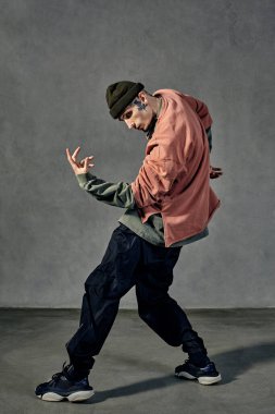 Dövmeli, yüzlü, küpeli, sakallı genç yakışıklı adam. Şapka, günlük kıyafetler ve siyah spor ayakkabılar giymişti. Gri arka planda dans etmek. Dans salonu, hip-hop. Tam uzunluk, boşluğu kopyala