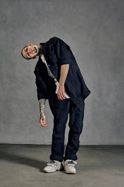 Dövmeli, küpeli, sakallı esnek bir adam. Beyaz tişört, spor ayakkabı, siyah kot gömlek ve pantolon giymişti. Gri arka planda dans etmek. Dans salonu, hip-hop. Tam uzunluk, boşluğu kopyala