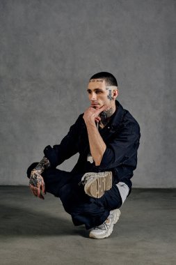 Dövmeli, küpeli, sakallı aktif bir sanatçı. Beyaz tişört, spor ayakkabı, siyah kot gömlek ve pantolon giymişti. Gri arka planda numaralar gösteriyor. Dans salonu, hip-hop. Kapat, boşluğu kopyala