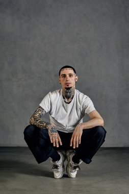 Dövmeli, yüzlü, küpeli, sakallı esnek bir sanatçı. Beyaz tişört ve spor ayakkabısı, siyah spor pantolon giymişti. Gri arka planda çorapların üzerine çömelmek. Dans salonu, hip-hop. Kapat.