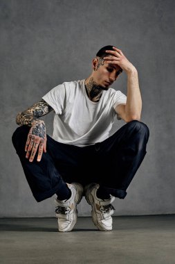 Dövmeli, yüzlü, küpeli, sakallı güçlü bir adam. Beyaz tişört ve spor ayakkabısı, siyah spor pantolon giymişti. Gri arka planda çorapların üzerine çömelmek. Dans salonu, hip-hop. Kapat.
