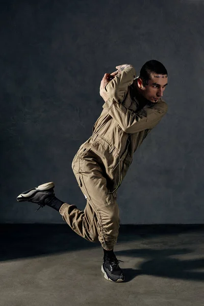Young handsome guy with tattooed body and face, earrings, beard. Dressed in khaki overalls and black sneakers. He is dancing against gray studio background. Dancehall, hip-hop. Full length, copy space
