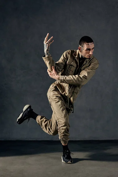 Young modern man with tattooed body and face, earrings, beard. Dressed in khaki overalls and black sneakers. He is dancing against gray studio background. Dancehall, hip-hop. Full length, copy space