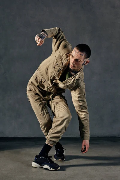 Young flexible guy with tattooed body and face, earrings, beard. Dressed in khaki overalls and black sneakers. He is dancing against gray studio background. Dancehall, hip-hop. Full length, copy space