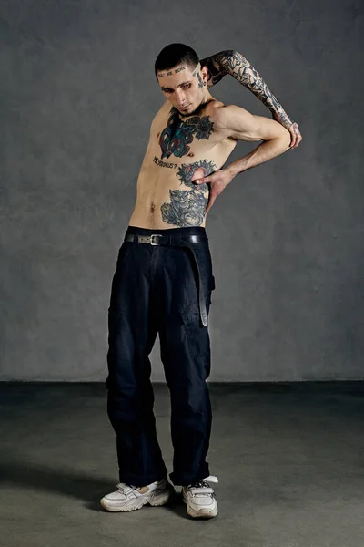 Flexible male with tattooed body and face, naked torso, beard. Dressed in black pants and white sneakers. He is dancing on gray studio background. Dancehall, hip-hop. Full length, copy space