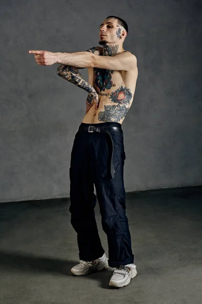 Flexible guy with tattooed body and face, naked torso, beard. Dressed in black pants and white sneakers. He is dancing against gray studio background. Dancehall, hip-hop. Full length, copy space
