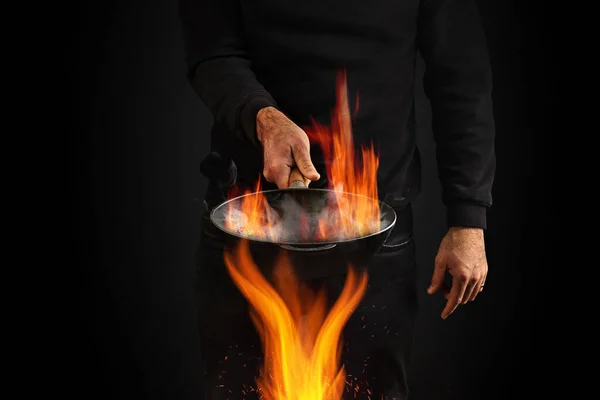 ジャンパーとジーンズを着た若い男 黒いスタジオを背景に 火の上で煙を上げて火鍋を焼いている 料理のコンセプト スペースをコピー — ストック写真