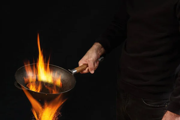 ジャンパーとジーンズを着た若い男 彼は黒いスタジオの背景を背景に火の上で焼き鍋を保持しています 料理のコンセプト スペースをコピーし サイドビュー — ストック写真
