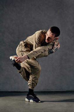 Dövmeli, yüzlü, küpeli, sakallı genç ve çekici bir adam. Haki rengi tulum ve siyah spor ayakkabı giymişti. Gri stüdyo arka planında dans ediyor. Dans salonu, hip-hop. Tam uzunluk, boşluğu kopyala