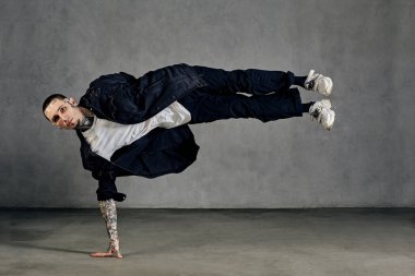 Dövmeli, yüzlü, küpeli, sakallı esnek bir adam. Beyaz tişört, spor ayakkabı, siyah kot gömlek ve pantolon giymişti. Gri arka planda numaralar yapıyor. Dans salonu, hip-hop. Tam uzunluk