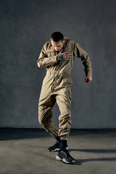 Young strong performer with tattooed body and face, earrings, beard. Dressed in khaki overalls and black sneakers. He is dancing against gray background. Dancehall, hip-hop. Full length, copy space