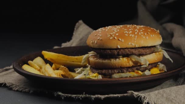 La caída de papas fritas en el plato oscuro con arpillera debajo de ella, junto a la hamburguesa fresca. Mesa negra y fondo. Comida rápida, servir. De cerca. — Vídeo de stock