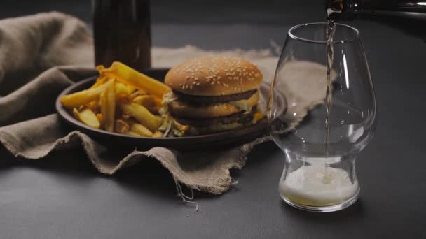 Pouring fresh and cold beer from dark bottle into glass with white foam on top. French fries on dark plate next to hamburger. Black table. Close up — Stock Video