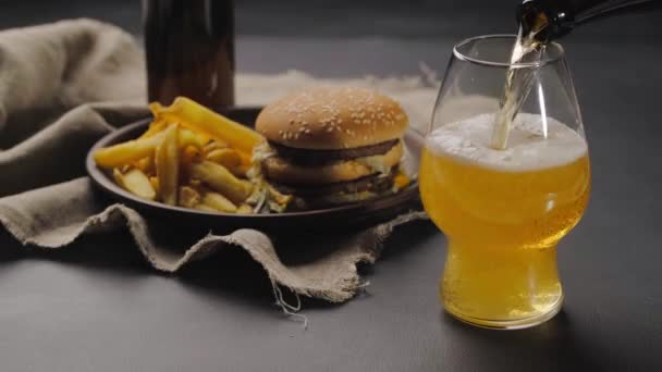 ダークボトルからグラスに新鮮なクラフトビールを注ぎ、上に白い泡が乗っています。ハンバーガーの横にある暗いプレートの上にフランスのフライドポテト。黒のテーブル。閉じろ! — ストック動画