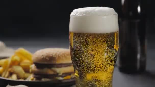 Пенящееся пиво в стакане с пузырьками, выходящими наружу, белая пена сверху. Размытый фон, картошка фри и гамбургер на темной тарелке. Фастфуд. Закрыть — стоковое видео