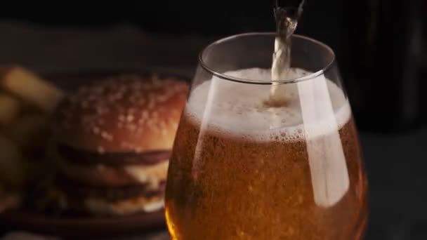 Verter cerveza artesanal en vidrio, espuma blanca en la parte superior. Fondo borroso con papas fritas y hamburguesa en el plato. Fondo negro. Comida rápida. De cerca. — Vídeo de stock