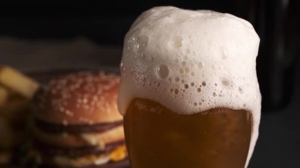 Pouring beer into glass with white overflowing foam slowly flowing down a glass. Blurred background with french fries and hamburger. Black background — Stock Video