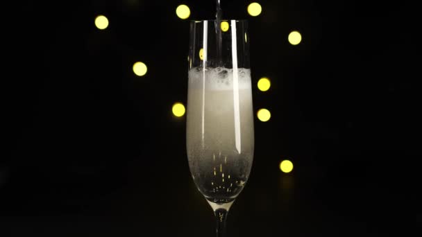 Verter champán o vino espumoso en una copa contra el bokeh de vacaciones de oro, fondo negro. Sale burbujas y espuma blanca en la parte superior. De cerca. — Vídeo de stock