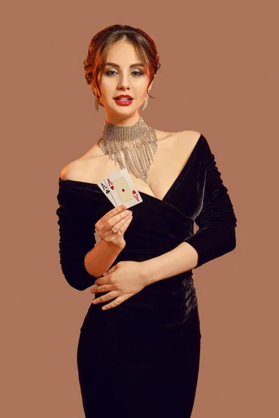 Chica morena, hombros desnudos, en vestido negro y joyas. Sonriendo, mostrando dos cartas, posando sobre fondo marrón. Poker, casino. Primer plano — Foto de Stock