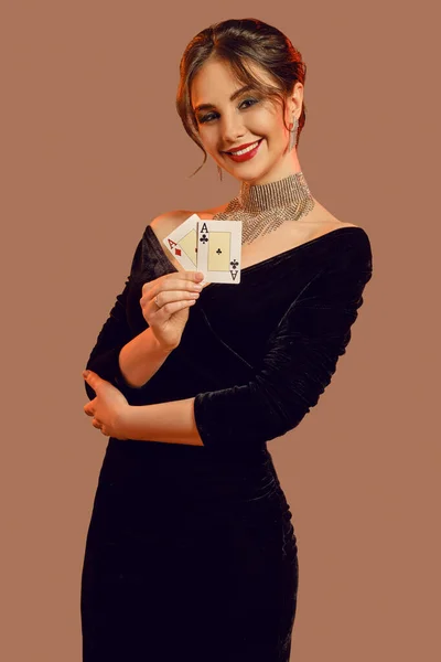 Brunette model, bare shoulders, in black dress and jewelry. Smiling, showing two playing cards, posing on brown background. Poker, casino. Close-up