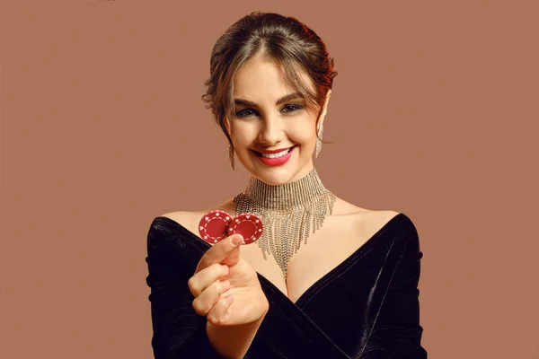 Modello bruna in abito nero e gioielli lucidi. Sorrise, mostrando due patatine rosse, in posa su sfondo marrone. Poker, casinò. Primo piano — Foto Stock