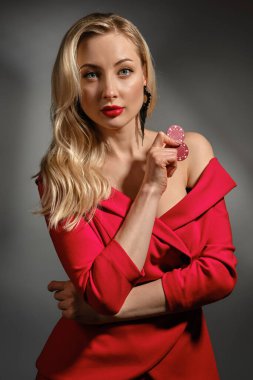 Çıplak omuzlu, kırmızı elbiseli ve siyah küpeli sarışın kadın. Gri arka planda poz veren iki çip gösteriyor. Poker, kumarhane. Yakın plan.