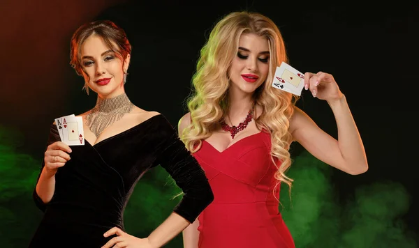 赤と黒のドレス ジュエリーで 明るく化粧をした2人の美しい女性 彼らは笑みを浮かべて トランプを示す 煙のようなカラフルな背景に対してポーズ ギャンブル ポーカー カジノ コピースペース — ストック写真