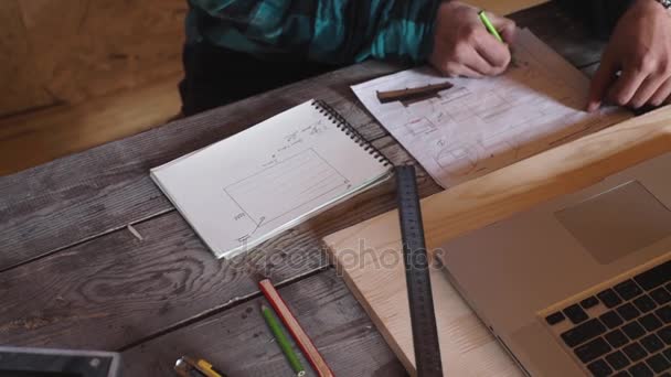 Деревообработчик работает в своей мастерской с оборудованием для рисования, бумагой, линейкой, карандашом и ноутбуком — стоковое видео