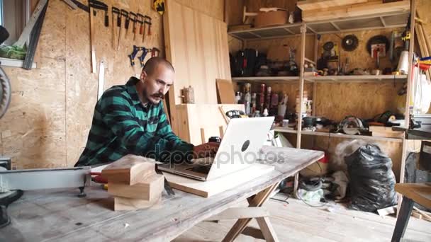 Retrato de bigote negrita carpintero retirado sentado en su taller y trabajando con el ordenador portátil. Pequeñas empresas . — Vídeo de stock