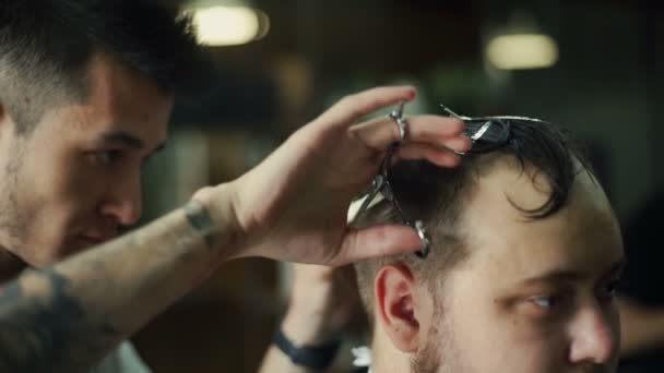 Young barber trimming mans hair with scissors in barbershop — Stock Video