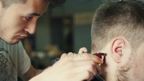 Corte perfeito. Visão traseira close-up de homem barbudo jovem recebendo corte de cabelo por cabeleireiro com lâmina elétrica enquanto sentado na cadeira na barbearia — Vídeo de Stock