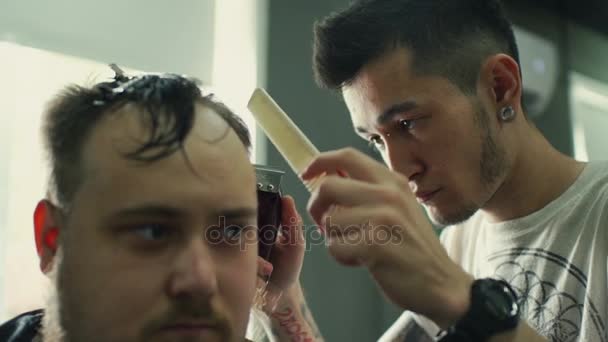 Corte perfeito. Visão traseira close-up de homem barbudo jovem recebendo corte de cabelo por cabeleireiro com lâmina elétrica enquanto sentado na cadeira na barbearia — Vídeo de Stock