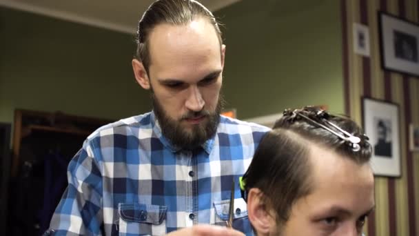 Atractivo peluquero joven está cortando el cabello humano con las tijeras. Está mirando el cabello con concentración. El barbudo levanta la barbilla. — Vídeos de Stock