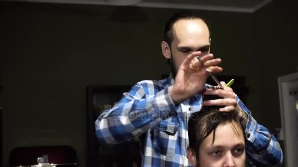 Barbeiro jovem atraente está cortando cabelo humano com a tesoura. Ele está olhando para o cabelo com concentração. O homem barbudo está levantando o queixo — Vídeo de Stock