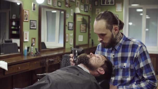 Homem barbudo atraente recebendo sua barba raspada por barbeiro moderno na barbearia — Vídeo de Stock