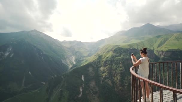 Mujer joven de pie en el balcón y tomando una foto de las montañas cercanas — Vídeo de stock