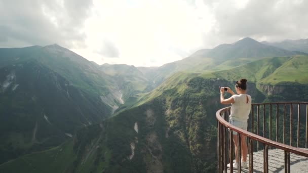 Mujer joven de pie en el balcón y tomando una foto de las montañas cercanas — Vídeos de Stock