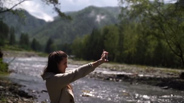 Portret van de jonge vrouw nemen mobiele foto selfie op de oever van de rivier van de berg in zonnige dag lacht — Stockvideo