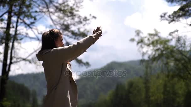 Retrato de una joven sonriente tomando foto autofoto móvil en la orilla del río de montaña en un día soleado — Vídeos de Stock