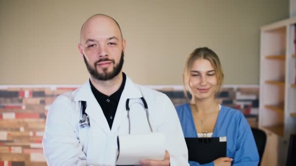 Adulto 30 homem médico masculino com uma enfermeira de 20 anos de uniforme estão posando no consultório médico 4k — Vídeo de Stock