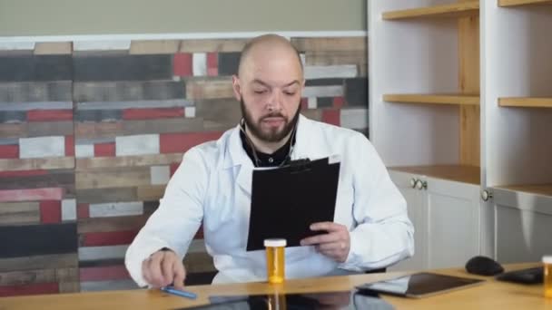 Portret dorosłych 30s łysy Brodaty lekarz siedzi na jego gabinet lekarski office wśród tabela 4k — Wideo stockowe